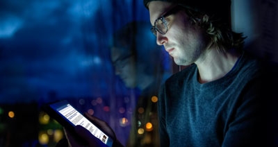 Man sitting against office window at dawn browsing iPad social media with Seattle city lights in background.